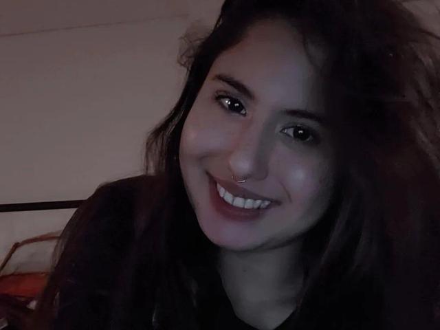 AuroraMonroe has dark, wavy hair, a nose ring, and a bright smile, wearing a black shirt, in dim lighting. Her tits are not visible.