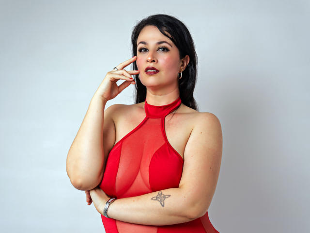 CarolinnaSaenz with dark hair, wearing a red sheer dress, touching her cheek, showing a star tattoo on her right forearm, and a silver bracelet.