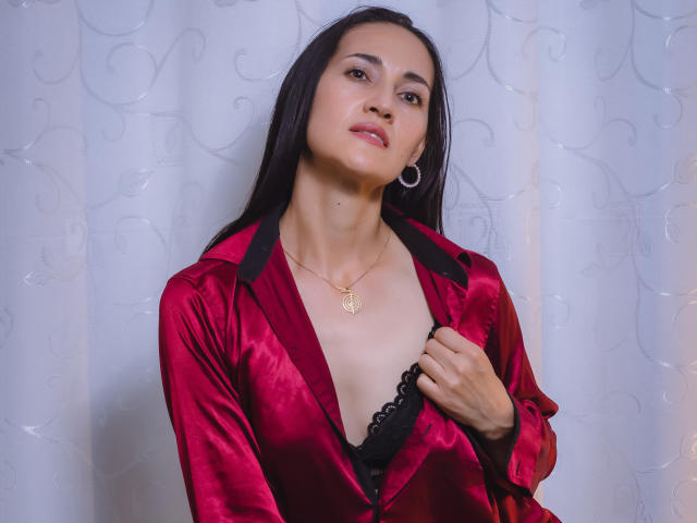 MiaMott with long black hair, wearing a red satin robe, exposing black lace bra, holding robe, small gold heart necklace, hoop earrings, and a slight smile.