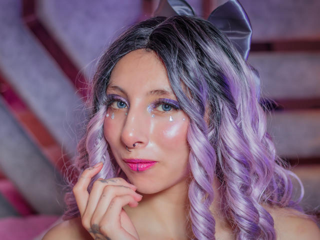 EssledthAnne with purple and silver curly hair, pink lipstick, nose ring, glitter on cheeks, and a black bow, holding her chin, looking at camera, with a cute, sexy expression.