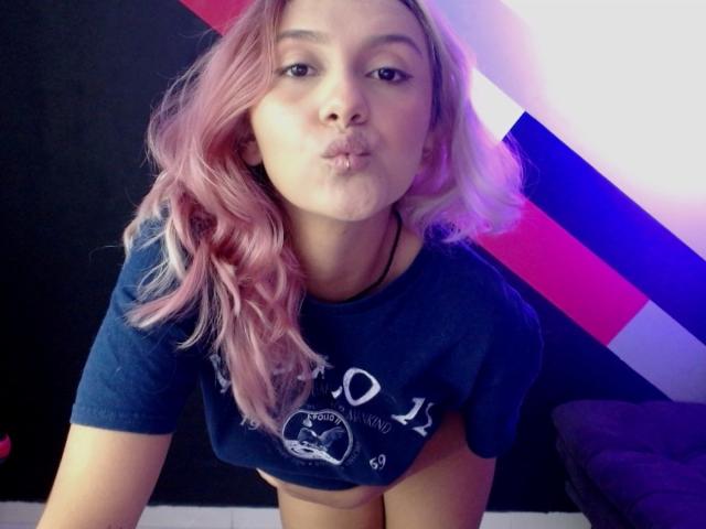 AlterEgoSecond with pink hair, pouty lips, and blue shirt, exposing her tits, leaning forward, wearing a black necklace, in a brightly colored room.