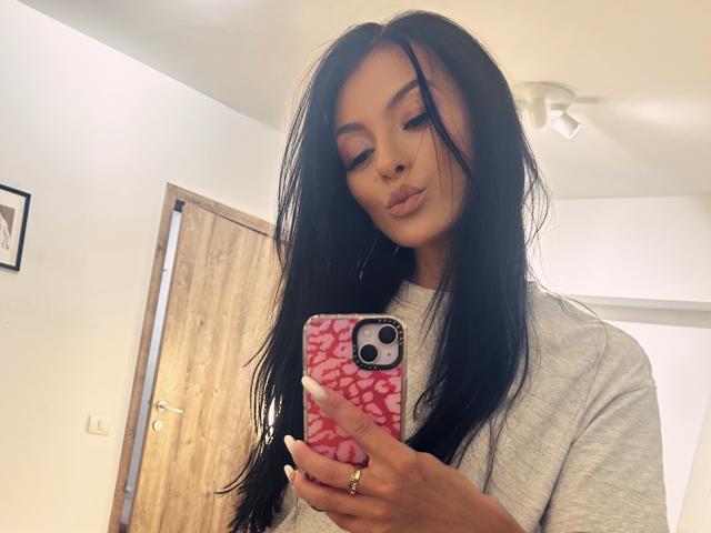 KinkyAmelie with long black hair, wearing a grey shirt, holding a pink phone with white patterns, looking down, wearing a gold ring on her finger.