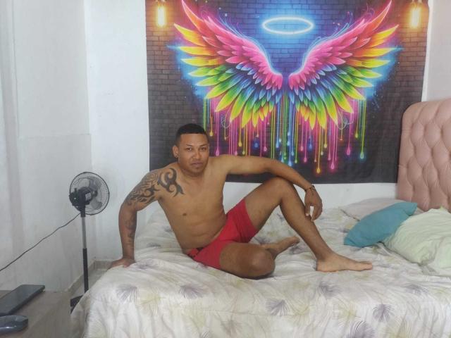 ChocoRamoHot, shirtless, red shorts, tattoo on right shoulder, sitting on bed, arms behind, legs crossed, electric fan on left, colorful wing wall art, pink tufted headboard, green pillow.