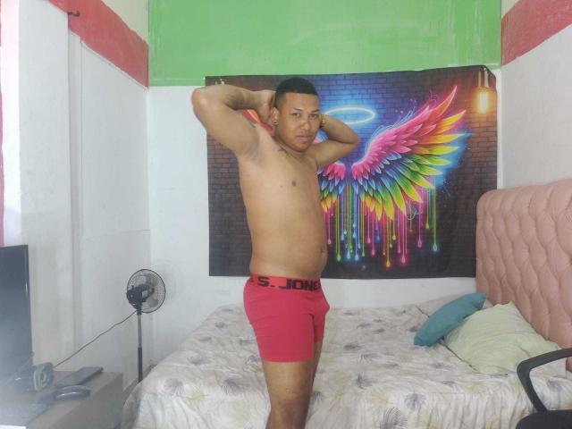 ChocoRamoHot stands shirtless in red boxer briefs, arms behind head, in a room with a pink bed and neon wing tapestry. He has a medium build and short hair.