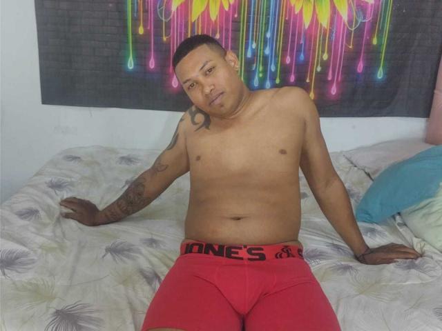 ChocoRamoHot, shirtless, sitting on bed, wearing red Jones underwear, tattooed arm, short black hair, medium brown skin, slightly chubby, looking at camera, fluorescent wall art behind.