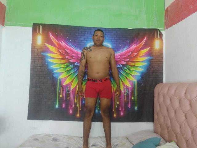 ChocoRamoHot stands in front of a colorful wing tapestry, wearing red boxer briefs, with a tattoo on his left shoulder, in a room with a pink quilted headboard.