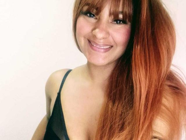 KarlysHoney with long red hair, smiling, wearing a black spaghetti strap top, showing her tits slightly, fair skin, and bangs covering her eyebrows.