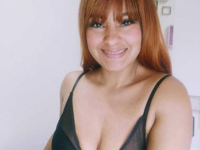 KarlysHoney has red hair, blue eyes, and a cute smile, wearing a black sheer top that reveals her small tits. She's looking at the camera, smiling.