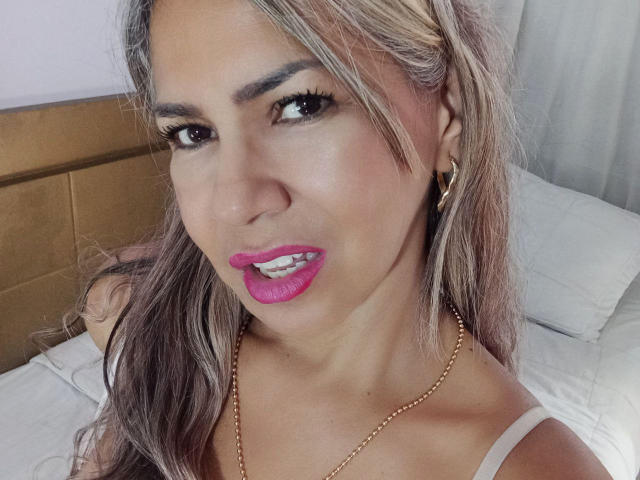 BrigiteWalsh with pink lipstick, gold hoop earrings, and a gold necklace, showing teeth, has long blonde and brown hair, wearing a white top, and brown eyes.