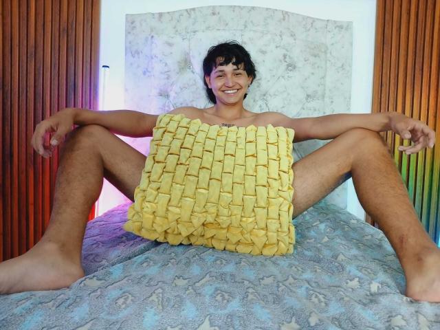 DanCristancho, naked with a yellow, woven, butt-cover, sitting on a bed with legs spread, smiling, short dark hair, tan skin, wood and marble background.