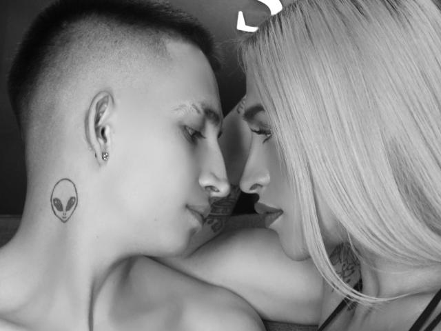 MyaAndJack69 with short, dark hair and a small alien tattoo on their neck, face close to MyaAndJack69 with blonde hair, both staring into each other's eyes.