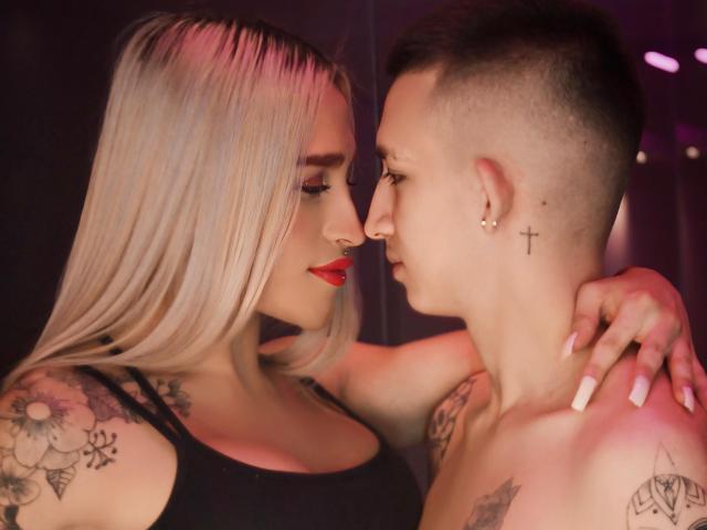 MyaAndJack69 with blonde hair and red lipstick, facing a shirtless MyaAndJack69 with a cross tattoo and short buzzcut, hands touching, tattoos visible, intimate, pinkish lighting.