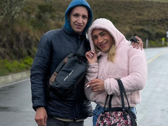 MyaAndJack69 with a cute ass, wearing a pink fuzzy hoodie, stands next to Jack69 in a blue hoodie, holding a camera and black bag, smiling on a road.