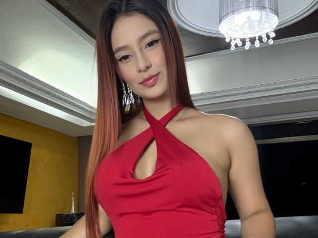 MyaSpecter with long brown hair, red halter dress showing small cleavage, and pink lips, standing in a room with a chandelier, looking at the camera.