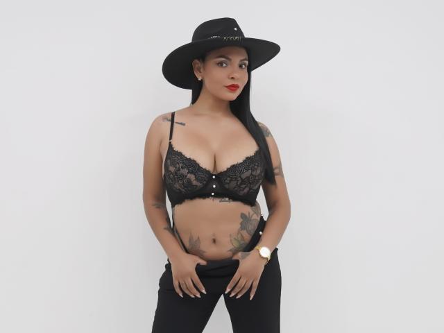 JessicaFiore has dark skin, long black hair, and red lipstick. She's wearing a black lace bra, black hat, and black pants, with tattoos on her arms and stomach.