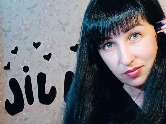 Jill Silvies, con cabello negro largo, ojos azules y labios rojos, vestida con una blusa negra, mirando a la cámara con una ligera sonrisa. Sus pequeños senos son parcialmente visibles..