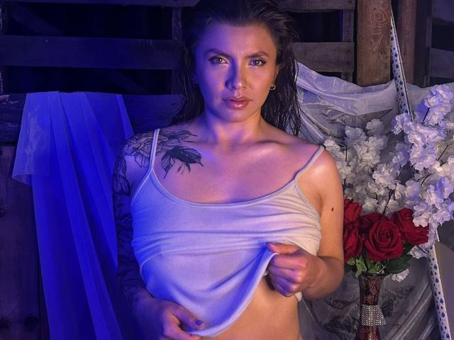 KarlaMorriss with wet hair, blue eyes, and a nose ring, lifting a white tank top to reveal her small tits and a black tattoo on her right shoulder.