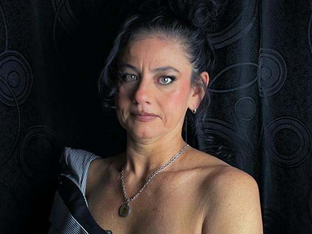 SasiLuna hat dunkles Haar, das zu einem unordentlichen Dutt gebunden ist, grüne Augen, eine kleine Nase und eine Kette mit einem grünen Anhänger. Sie trägt keine Oberteile, sondern eine schwarz-weiß gestreifte Schulterpartie..