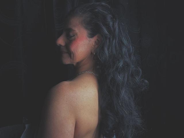 SasiLuna with long, wavy black hair, bare shoulders, and a pink blush on her cheek, wearing a silver necklace, standing in a dark, moody setting.