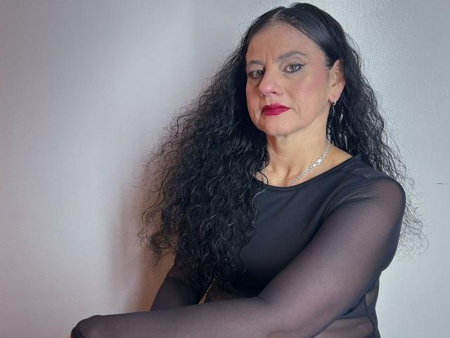 SasiLuna, com longos cabelos cacheados e pretos, vestindo uma blusa preta transparente, batom vermelho e brincos de prata, tem uma expressão confiante e os braços cruzados..