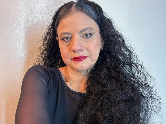 SasiLuna has long curly black hair, wearing a black sheer top, with red lipstick and a serious expression. She has a silver necklace and is standing against a white wall.
