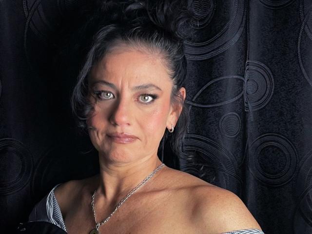 SasiLuna tiene el cabello oscuro recogido en un moño despeinado, ojos verdes y un rostro con labios fruncidos y arrugas visibles. Lleva un collar y un vestido con un hombro descubierto, mostrando sus pechos desnudos..