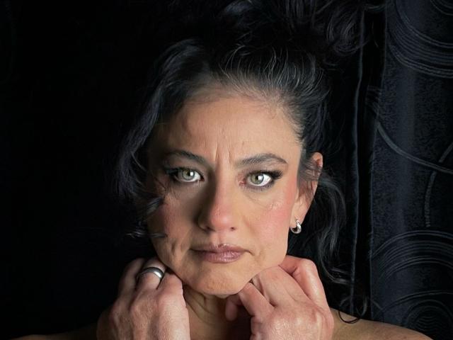 SasiLuna a les cheveux noirs et ondulés, et elle regarde vers le haut avec ses yeux verts. Elle se tient le menton avec les deux mains, portant une bague et des boucles d'oreilles de perles, sur un fond noir..