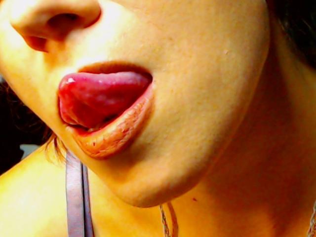 JuliaIce's tongue is out, wet and red, with her lips slightly parted. She's wearing a brown tank top, and her face is close to the camera.