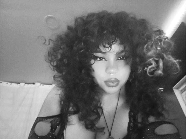 NeffellyRoss with curly dark hair, nose ring, and pouty lips, wearing a black mesh top, showing her large, bare tits. Black-and-white, gritty, selfie-style.