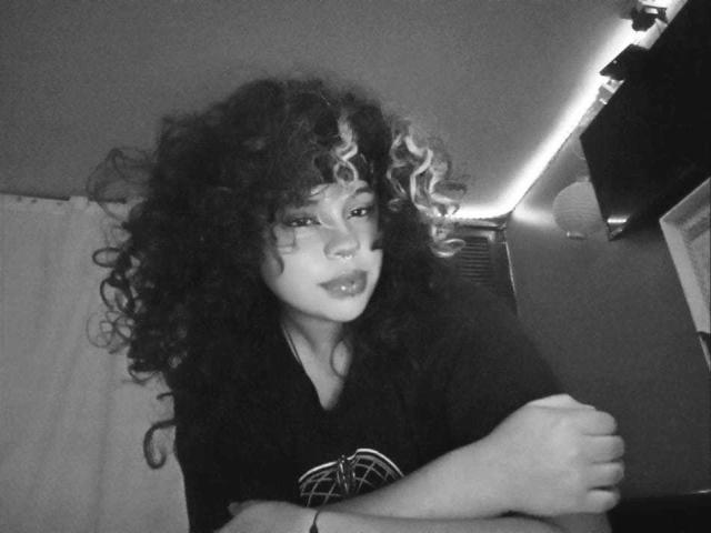 NeffellyRoss has curly dark hair with light blonde highlights, wearing a black shirt, sitting with arms crossed, looking slightly to the side in a black-and-white photo.