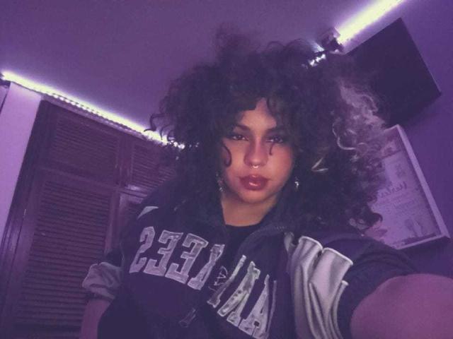 NeffellyRoss with curly dark hair, wearing a black and white sports jersey, purple lighting, nose ring, and red lipstick, taking a selfie. Her tits are slightly visible.