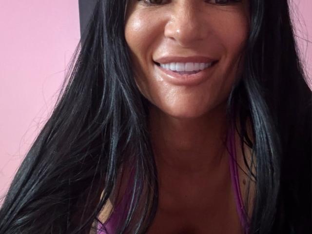AndraD has shiny brown skin, long black hair, and a big smile showing white teeth. She's wearing a pink top with visible cleavage. Her face is slightly sweaty.