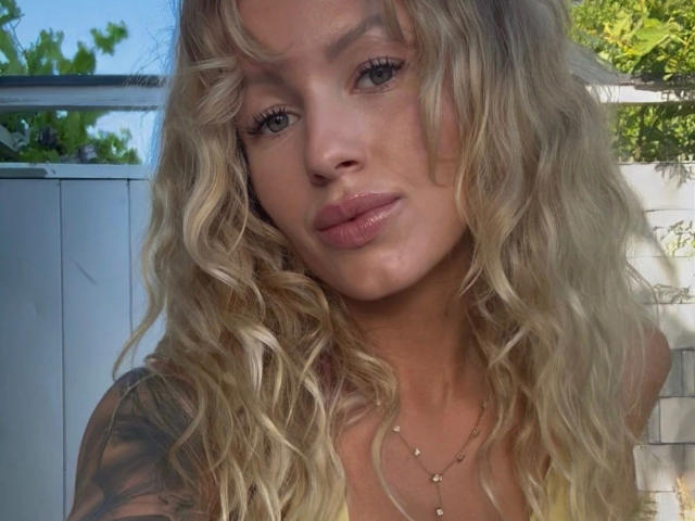 TinyTease-hot with wavy blonde hair, full pink lips, and a tattoo on her right arm, wearing a necklace, looking at the camera with a subtle smile.