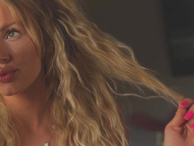 TinyTease-hot with wavy blonde hair, pink nails, and glossy red lips, holding a lock of hair, has green eyes and a slightly smiling face in a dimly lit setting.