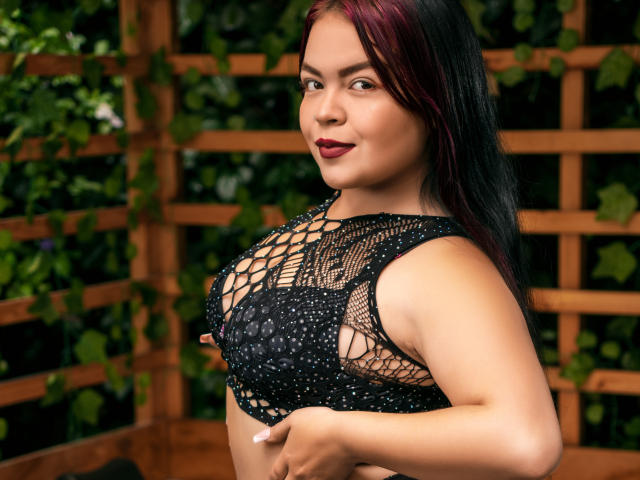 CharlottexEvans with dark red and black hair, wearing a black netted top, showing her big tits with visible nipples, standing with a confident smile, dark red lipstick.