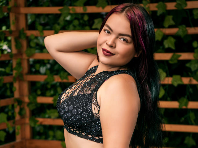 CharlottexEvans with dark red and black hair, wearing a black, lace-up, sleeveless top, showing her tits, arm raised, smiling with dark lipstick, against a wooden, leafy background.