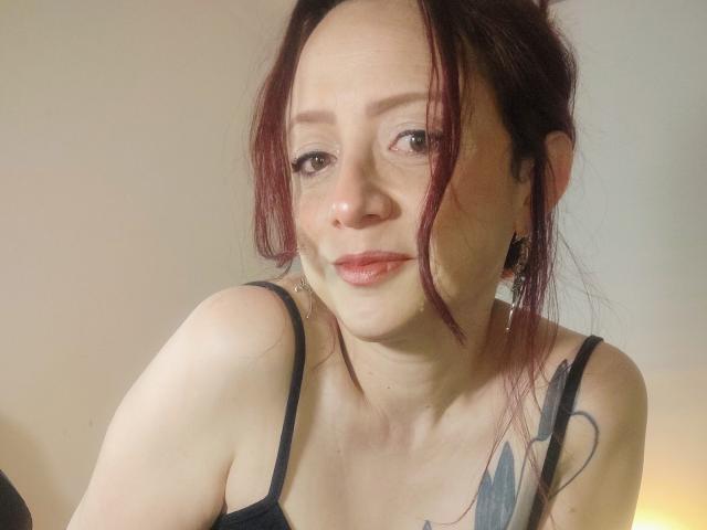 CatalinaMonyque has red hair, light skin, and subtle tattoos on her shoulder. She's wearing a black spaghetti strap top, smiling with slightly parted lips, and has earrings.