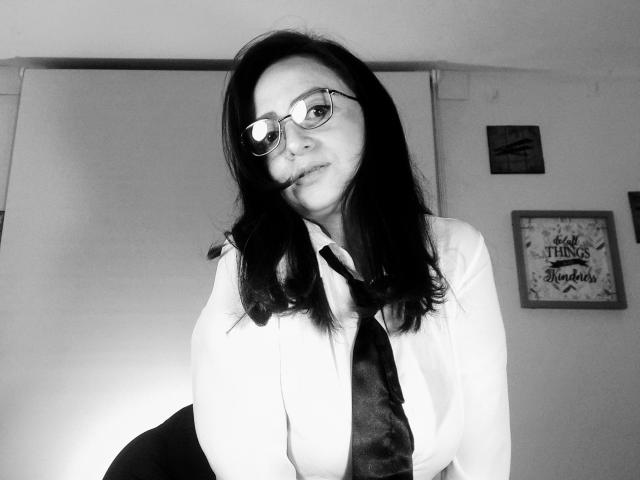 CatalinaMonyque with glasses, dark straight hair, white shirt, and black tie, leaning forward, smiling, her small tits visible, black skirt, grayscale photo.