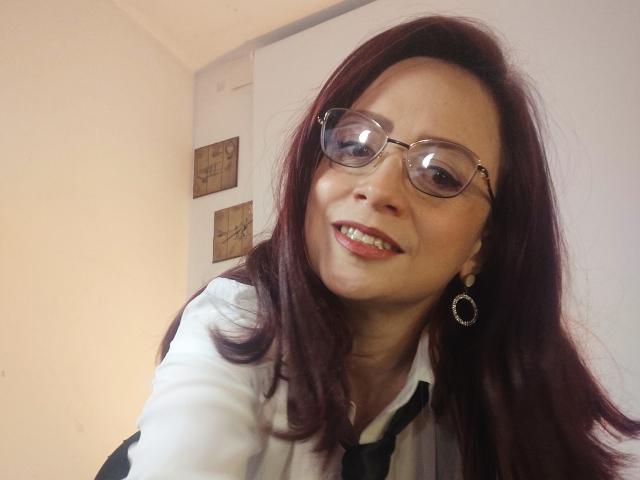 CatalinaMonyque has red hair, glasses, and is smiling with pink lips, wearing a white shirt and black tie, showing off her earrings and looking sexy.