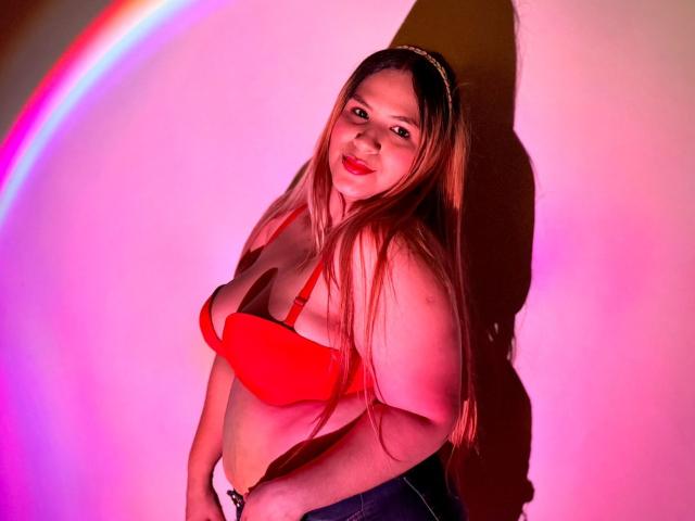 BrendaMarxhal with long brown hair, red bra showing her tits, pinkish-purple light, smiling, hands on hips, shadow on wall, wearing a gold headband, in a sexy pose.