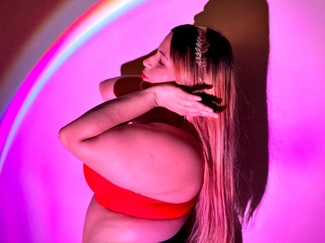 BrendaMarxhal with long blonde hair, red bra, and braid, touching her head, showing her ass, in a pink-lit, rainbow-circular-bokeh photo. Her arms are crossed.