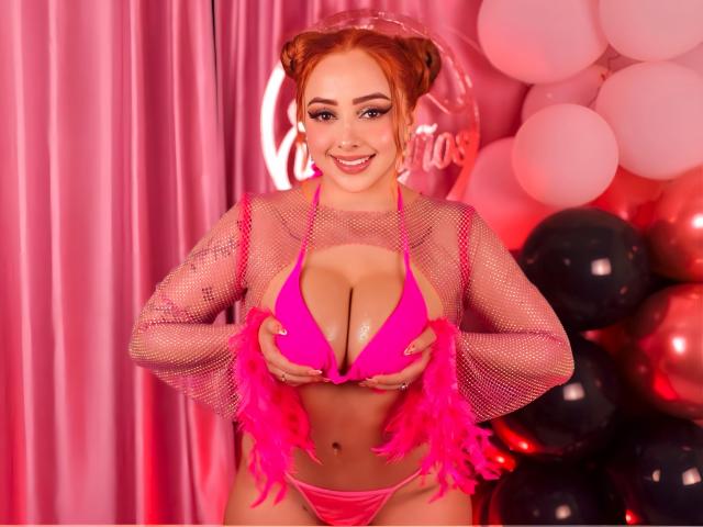 KylieCharm con i capelli rossi, che indossa una maglia di rete rosa, polsini di piume rosa e un bikini rosa acceso, tiene le sue grandi tette, sorridendo, in una stanza piena di palloncini rosa e rossi..