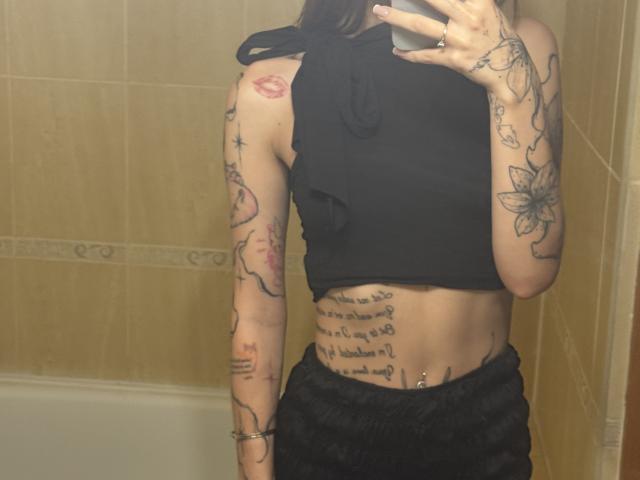 SensualMila has tattoos on her arms, a lip tattoo on her shoulder, a script tattoo on her belly, and a flower tattoo on her forearm. She's wearing a black top.