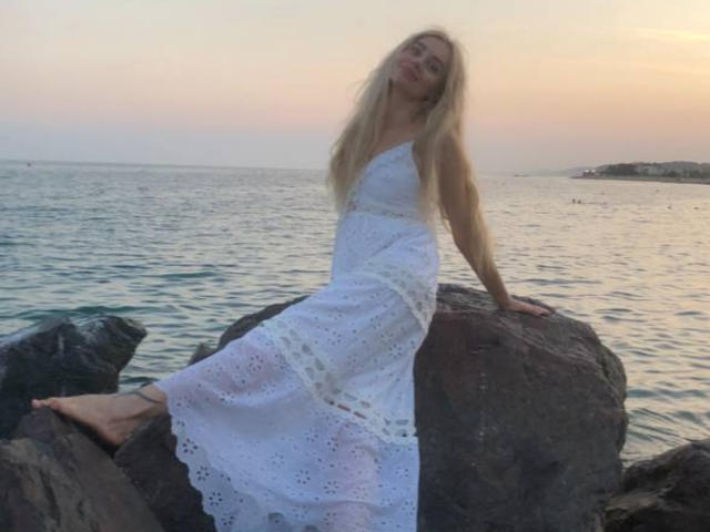 SabrinaSharm with long blonde hair, wearing a white, lace, sleeveless dress, sitting on rocks, her bare feet in the water, her ass slightly raised, at sunset.