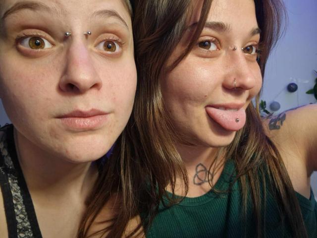 Two AliensForTwenty with piercings in their noses and eyebrows, one sticking out her tongue, both showing tattoos and wearing green and black tops.
