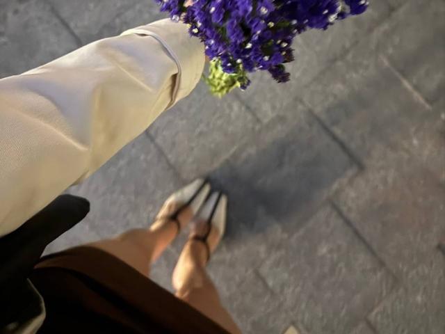 Le gambe di GingerFairy, indossate pantaloni beige, che tiene un bouquet di fiori viola, in piedi su un pavimento di pietra grigia con scarpe nere e bianche a punta..