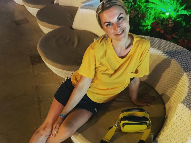 BelleLisaG, cabelo loiro curto, camisa amarela, shorts pretos, sentada em uma cadeira de vime curvada, sorrindo, bolsa amarela no assento, planta verde ao fundo, pernas descobertas, ambiente casual noturno..