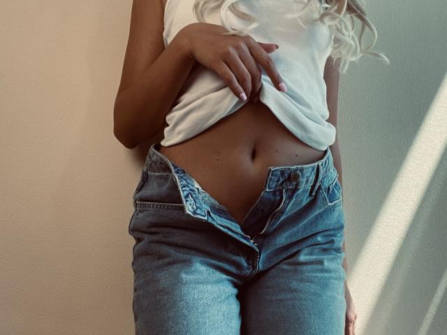 PrimeS has a flat, toned belly, wearing unbuttoned blue jeans, with a white shirt lifted to expose her navel, and has her hand on the shirt.