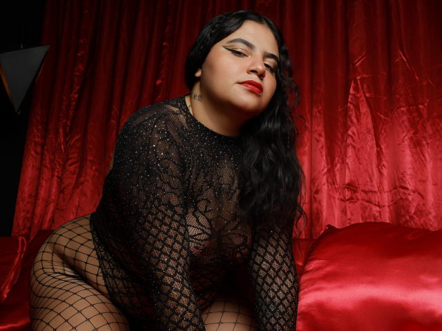 JannaFrank has black wavy hair, red lipstick, and wears a see-through black lace top with fishnet bottoms, showing her tits and pussy, sitting on red satin.