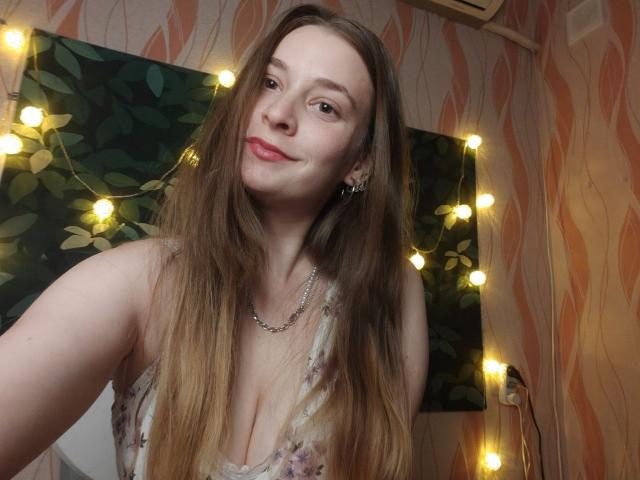 LunaBliss69 with long brown hair, smiling, wearing a floral dress with a low neckline, showing her tits, wearing a chain necklace, and fairy lights in the background.